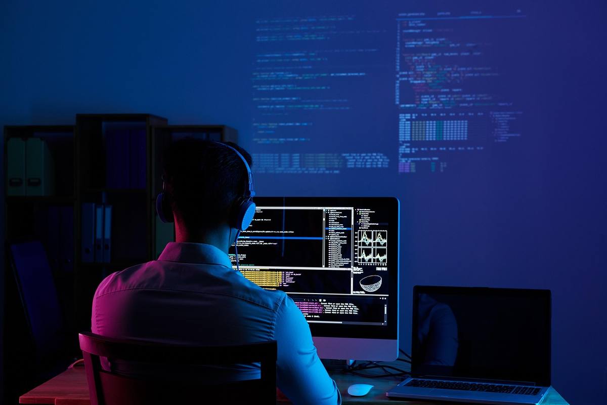 A developer in headphones working at a monitor full of code in a dark, blue-lit room